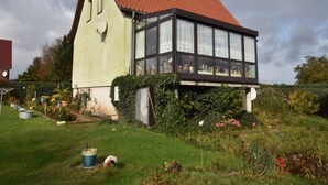 A traditional house with a red-tiled roof, a large windowed front section, and a garden with various plants and flowers. - Ferienzimmer in Kropelin (Kroepelin)