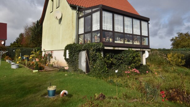 A traditional house with a red-tiled roof, a large windowed front section, and a garden with various plants and flowers. - Ferienzimmer in Kropelin (Kroepelin)