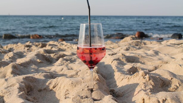 A glass of red drink with a straw on a sandy beach with the ocean in the background. - Ferienzimmer in Kropelin (Kroepelin)