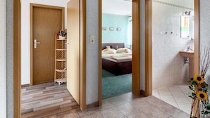 Een hotelkamer met een houten deur, een klein houten plankje met planten, een bed met een blauwe kap en een badkamer met een wastafel en spiegel.