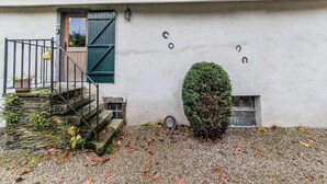 A building with a white exterior, a wooden door, a small staircase, a metal railing, a potted plant, a hedge, and a gravel ground. - Characteristic Holiday Home With Balcony (Gouvy)