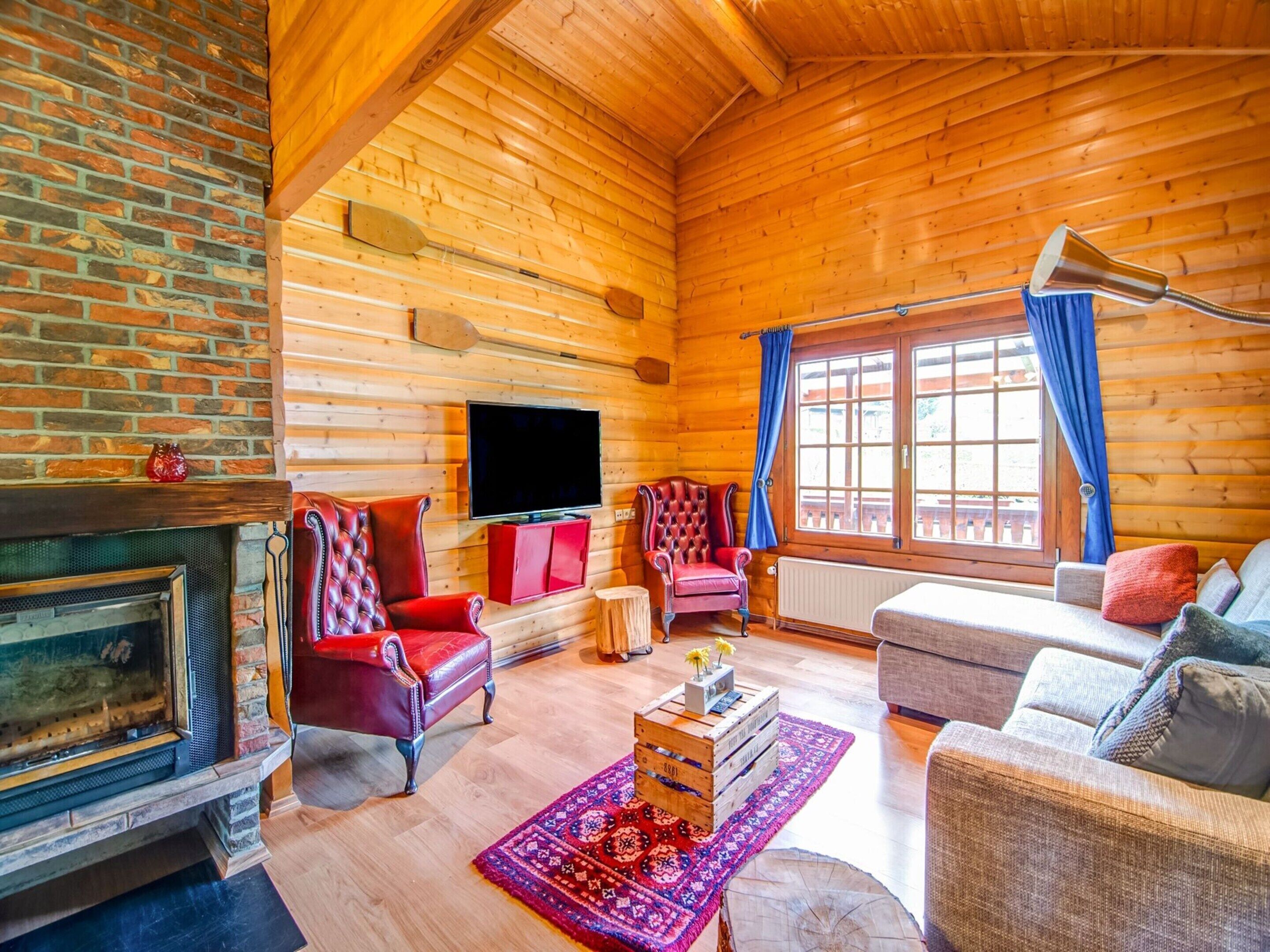 Interior acogedor de una cabaña de troncos con chimenea, televisor, un sillón rojo, un sofá y un ventanal con cortinas azules.