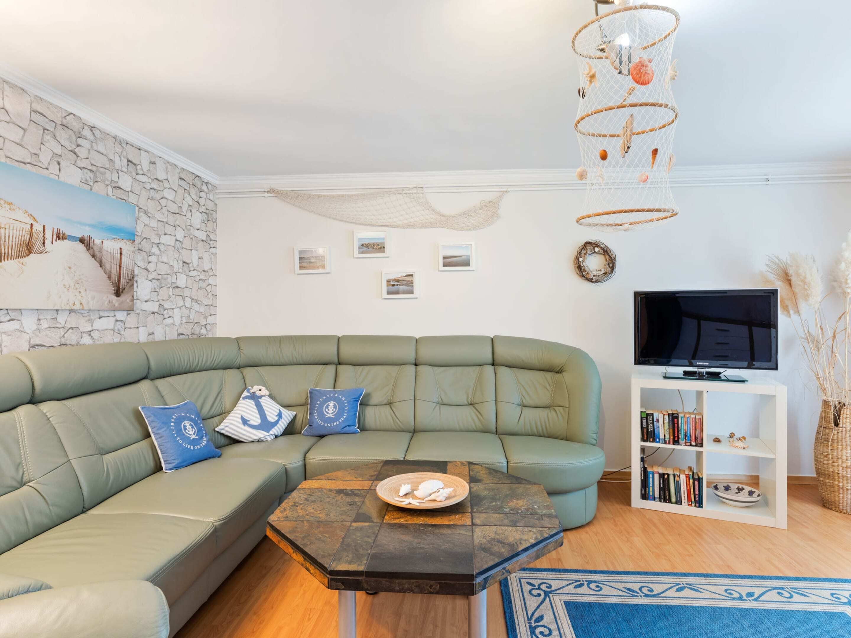 Una sala de estar moderna con un sofá seccional verde, una pared de piedra, un cuadro con temática de playa, una lámpara colgante y una estantería con libros y objetos decorativos.