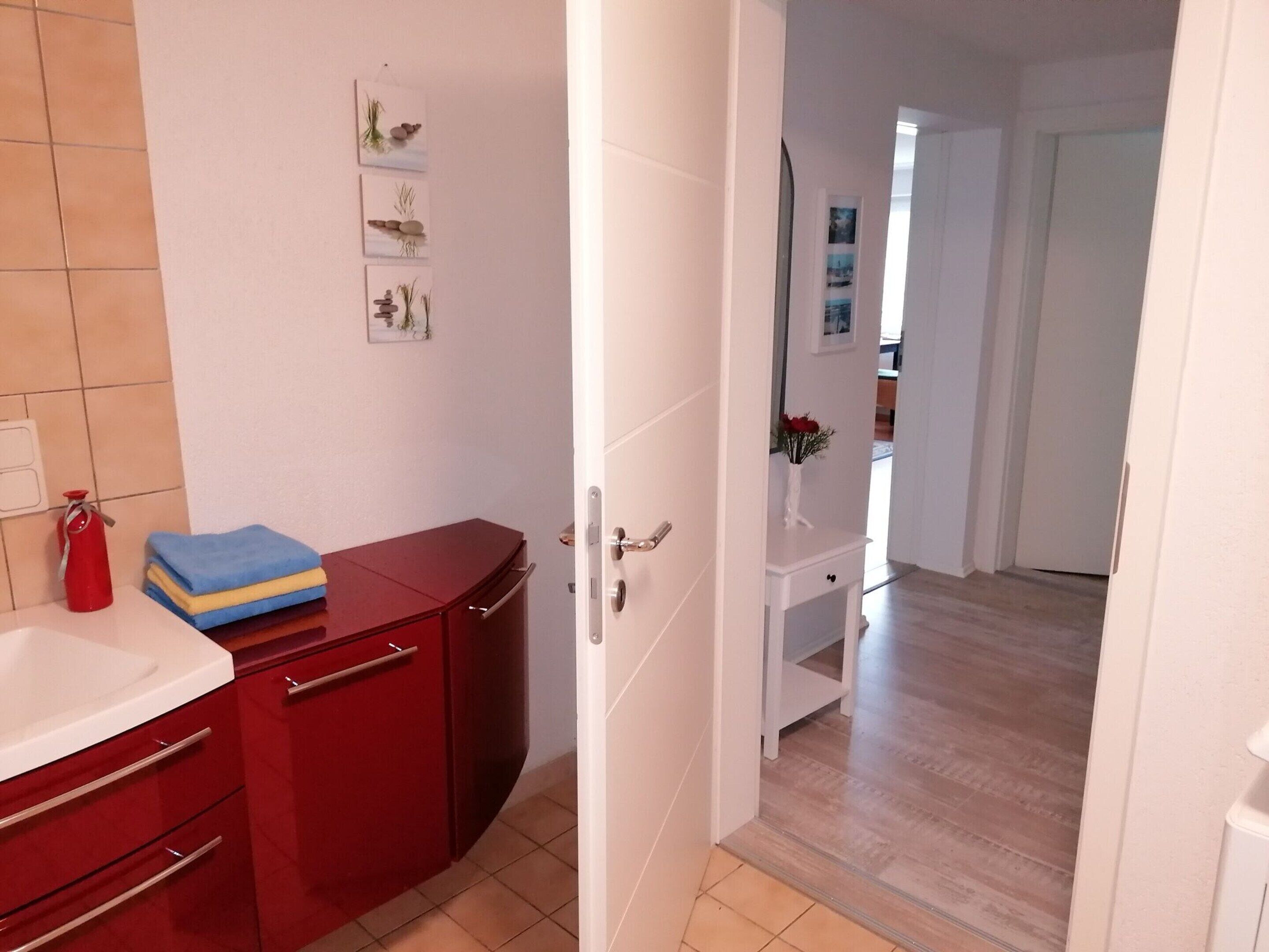 Une salle de bain avec un meuble rouge, un lavabo blanc et un extincteur rouge. Trois tableaux encadrés sont accrochés au mur.