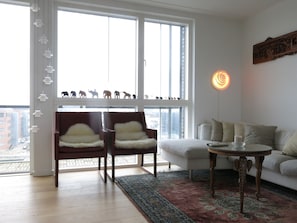 A modern living room with a large window, two armchairs, a sofa, a patterned rug, and a decorative wall piece. - Penthouse in Sydhavn 1135-1 (Copenhagen)