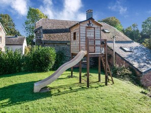 A wooden playhouse with a slide, a brick house, and a stone wall in the background.