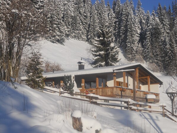 A wooden cabin with a snow-covered roof, surrounded by snow-laden trees and a clear sky.