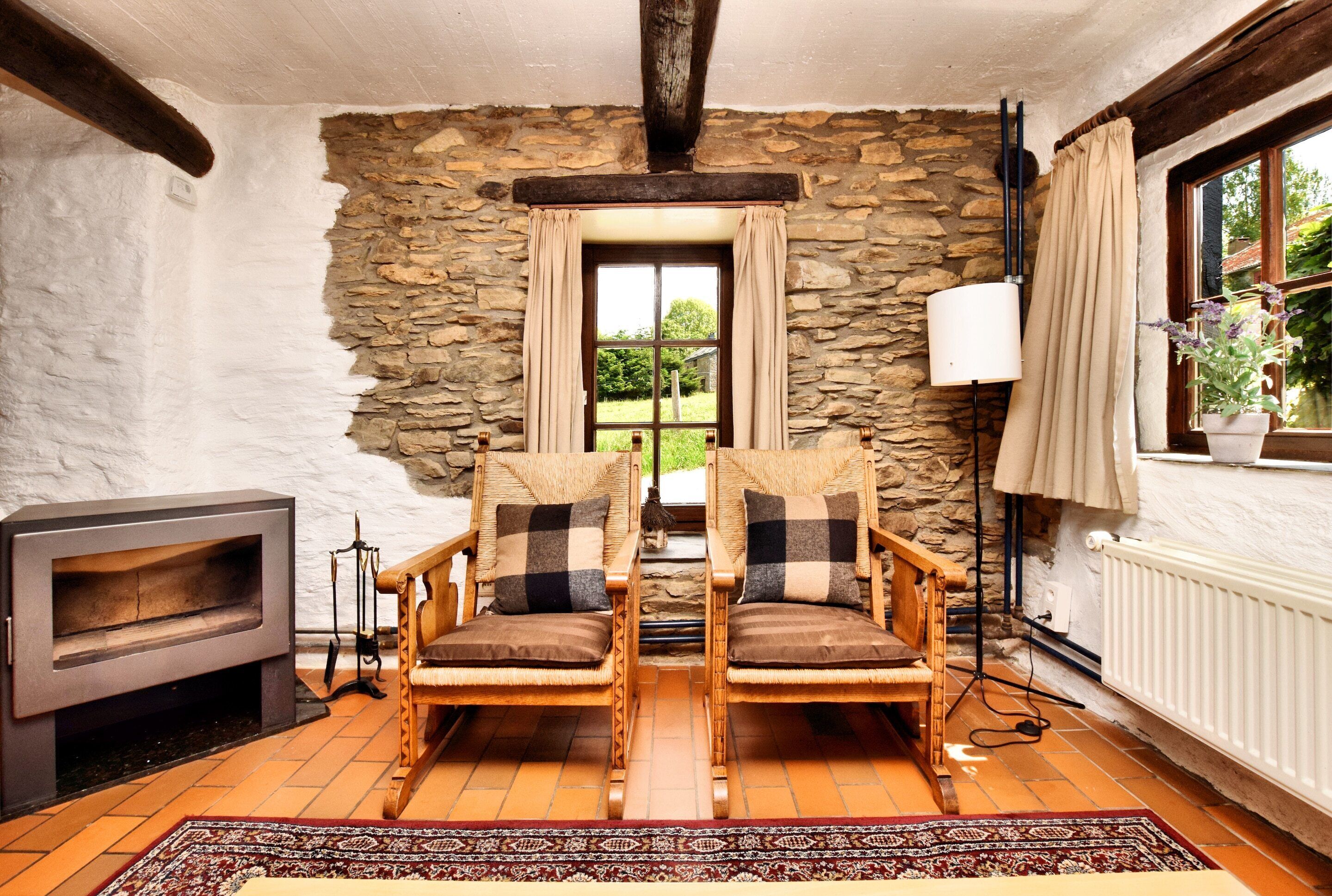 A cozy room with stone walls, a fireplace, two wooden chairs with cushions, a floor lamp, and a view of greenery outside.