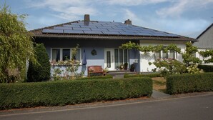 Une maison de plain-pied avec un toit équipé de panneaux solaires, un porche à l’avant avec un banc et un jardin bien entretenu.