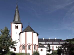 Una iglesia con un campanario alto y una torre redonda, rodeada de árboles y un edificio con numerosas ventanas.