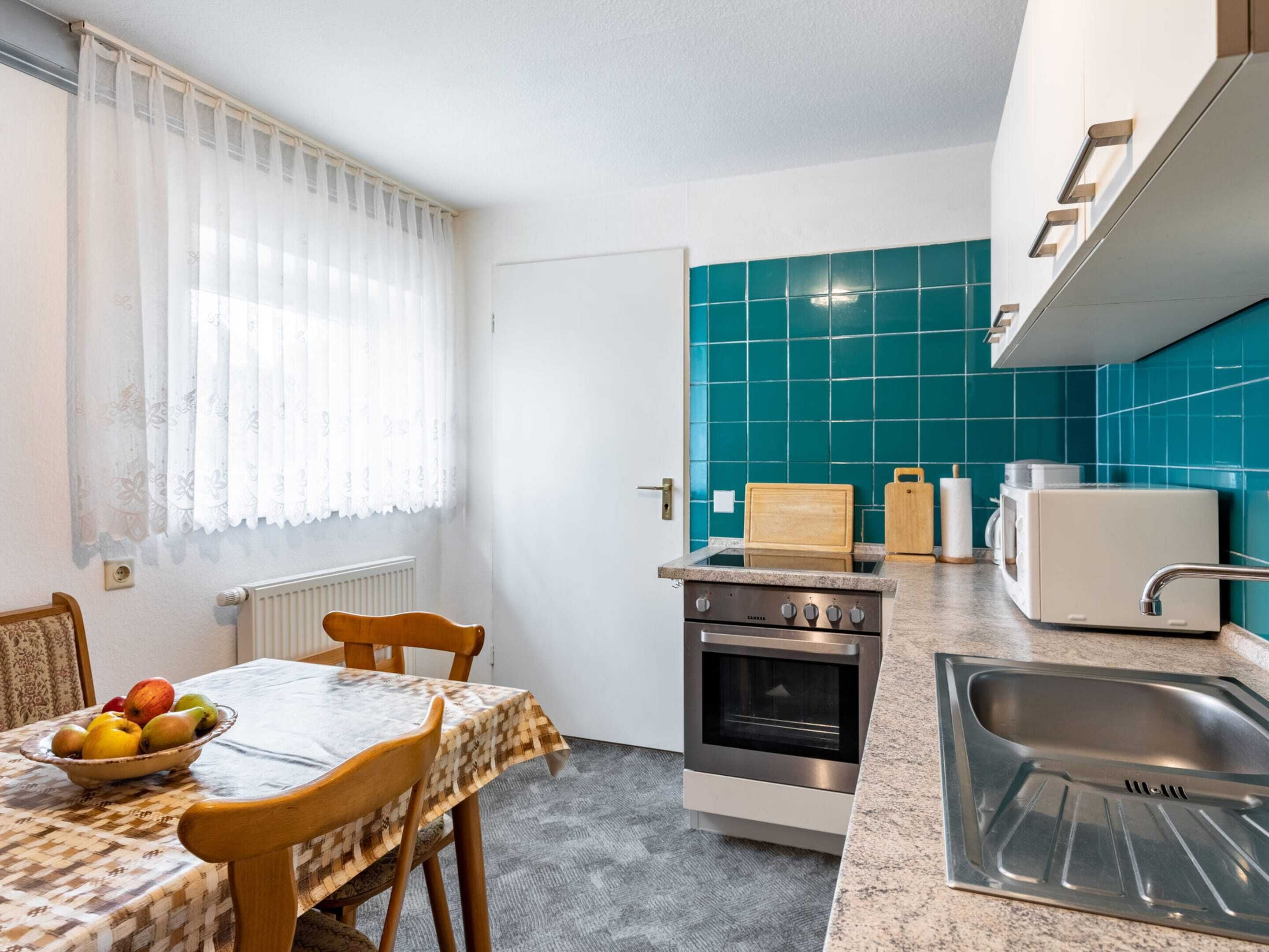 A compact kitchen with a stove, microwave, and sink. There is a table with a bowl of fruit and a chair.