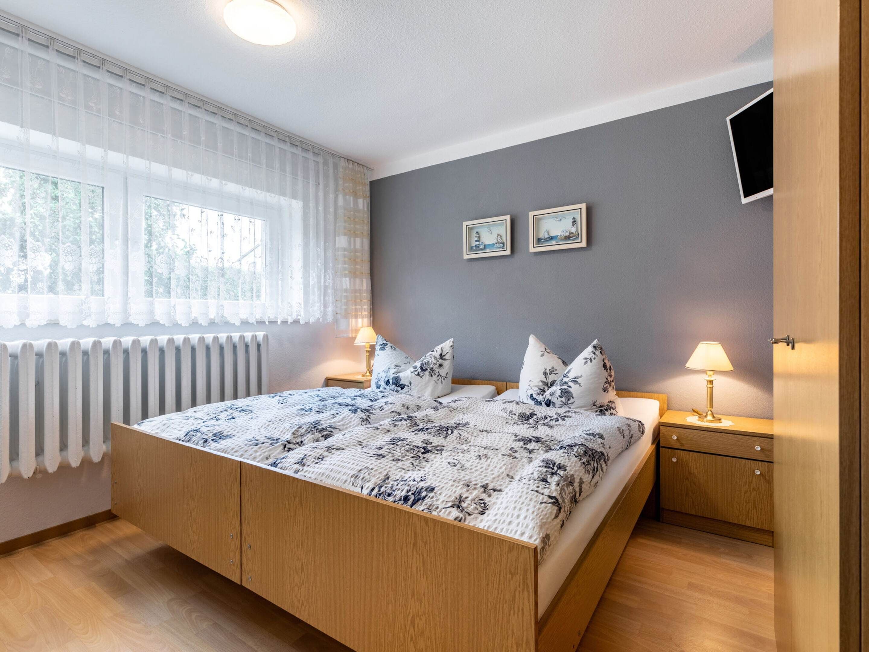 Un dormitorio con una cama de madera, dos mesitas de noche con lámparas, una ventana con cortinas translúcidas y dos cuadros en la pared.