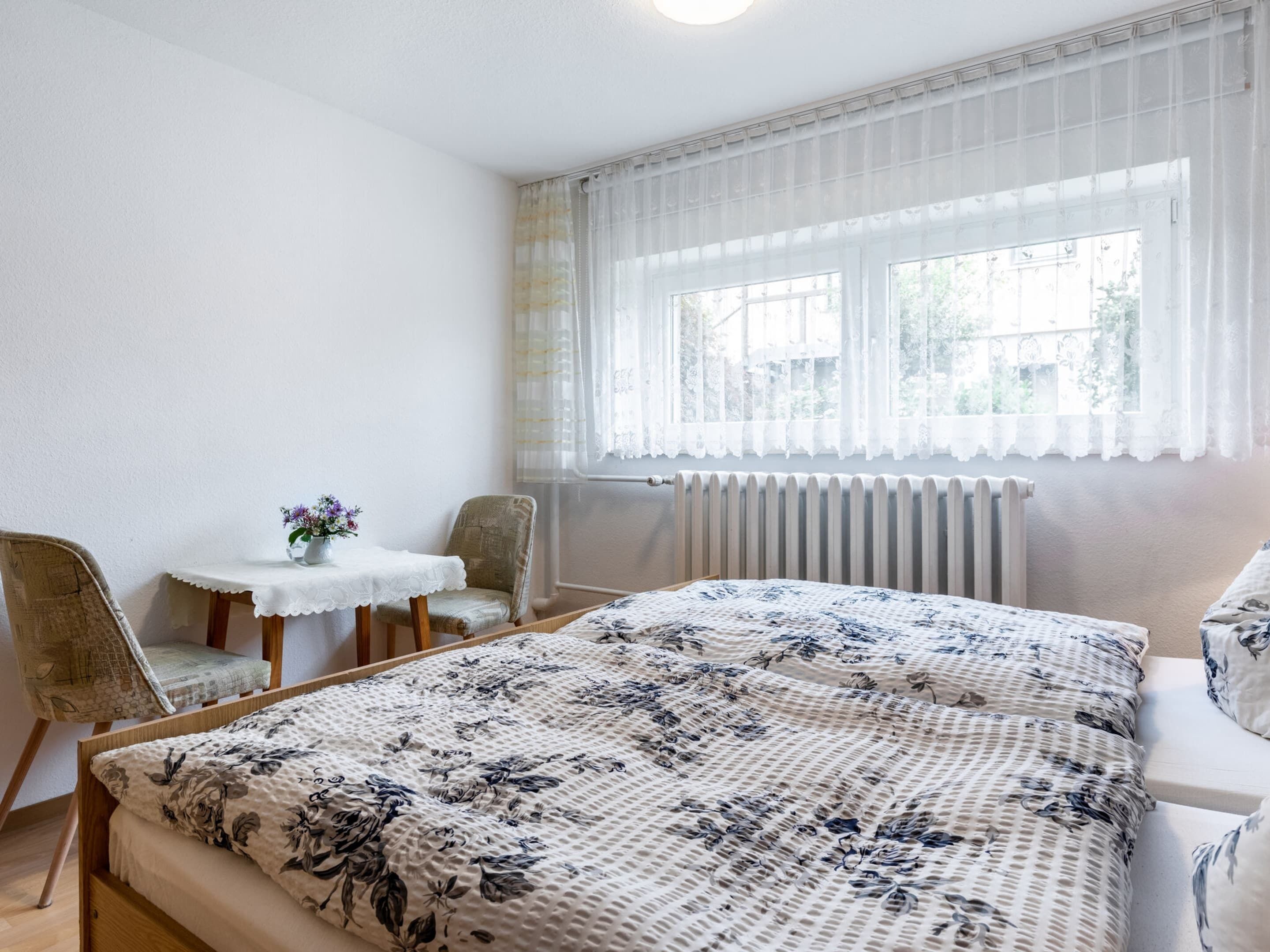 A bedroom with a bed, a chair, a small table with a vase of flowers, and a window with sheer curtains.