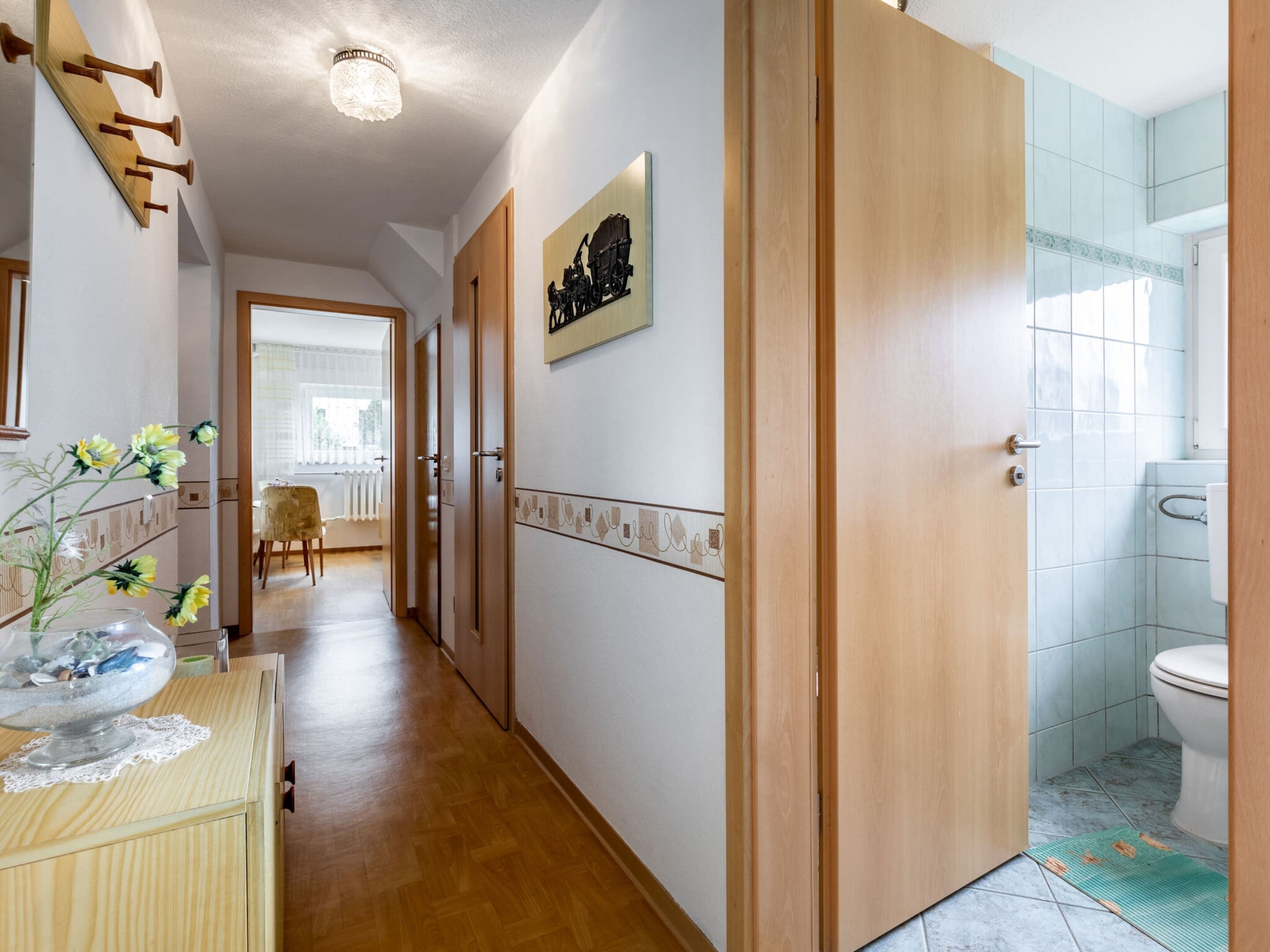 A hallway with wooden flooring, a wooden door, a bathroom with a toilet and tiled walls, and a dining area with a chair and table.