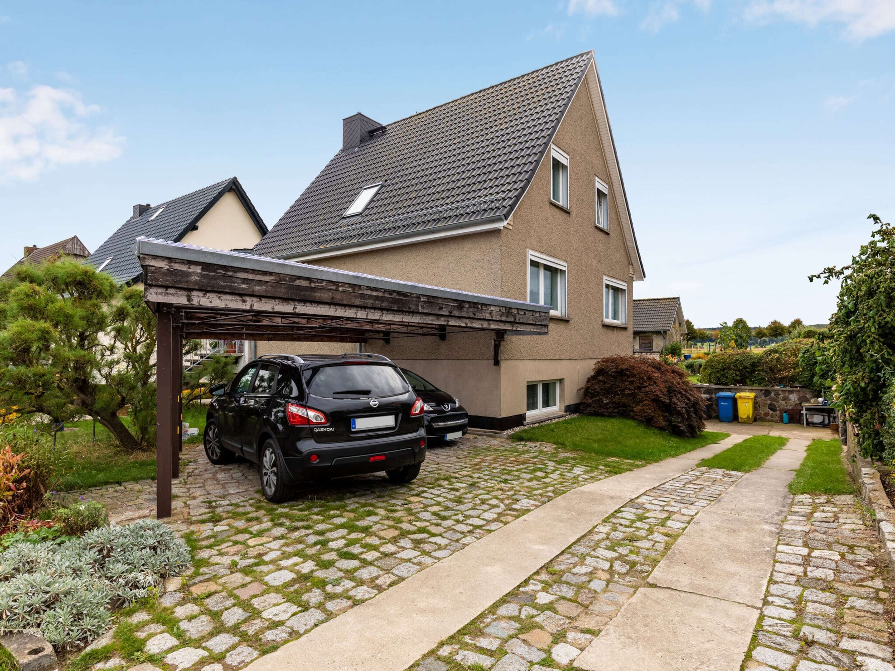 Ein Wohnhaus mit befestigter Auffahrt, überdachtem Carport und einem Garten mit Sträuchern und Rasen.