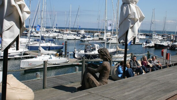 A marina with multiple sailboats docked, a statue of a lion, and people sitting on a wooden deck.