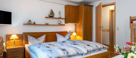A neatly arranged bedroom with a wooden bed, bedside tables, a mounted TV, and a shelf with decorative items.