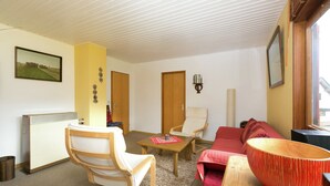 A living room with a wooden chair, a red sofa, a wooden table, and a red bowl. There are two doors, a radiator, and a framed picture on the wall. - Charming Holiday Home in Liebenscheid by Forest (Liebenscheid)