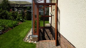 Une maison moderne avec une entrée dotée d’une porte en verre, un chemin en briques menant à la porte, et un jardin agrémenté de diverses plantes et d’une pelouse.