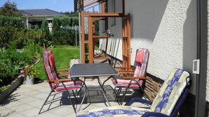 Une terrasse avec une table et quatre chaises, une pergola en bois et un jardin avec différentes espèces de plantes.