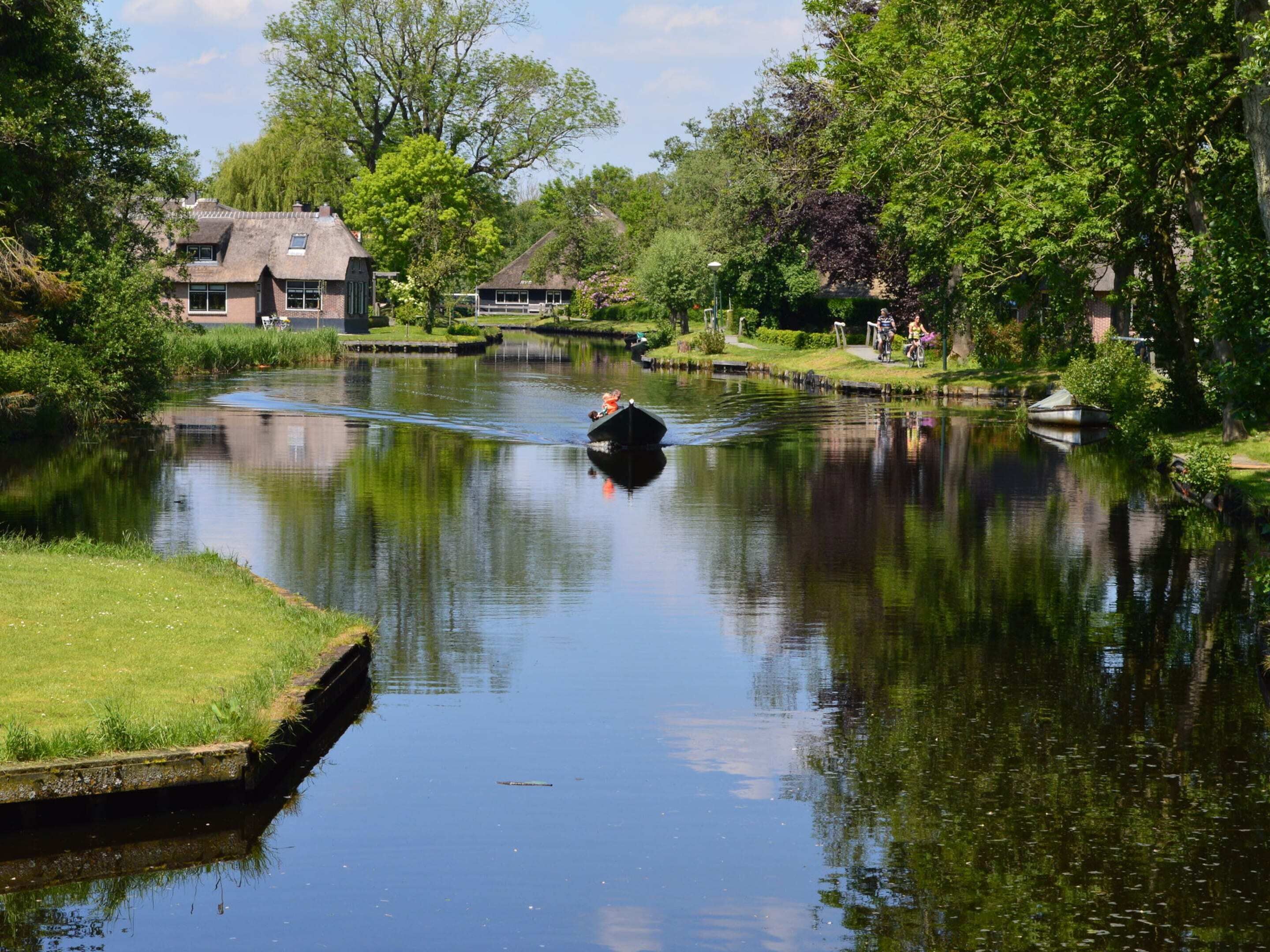 A calm river with a boat carrying two people, surrounded by lush greenery and houses.