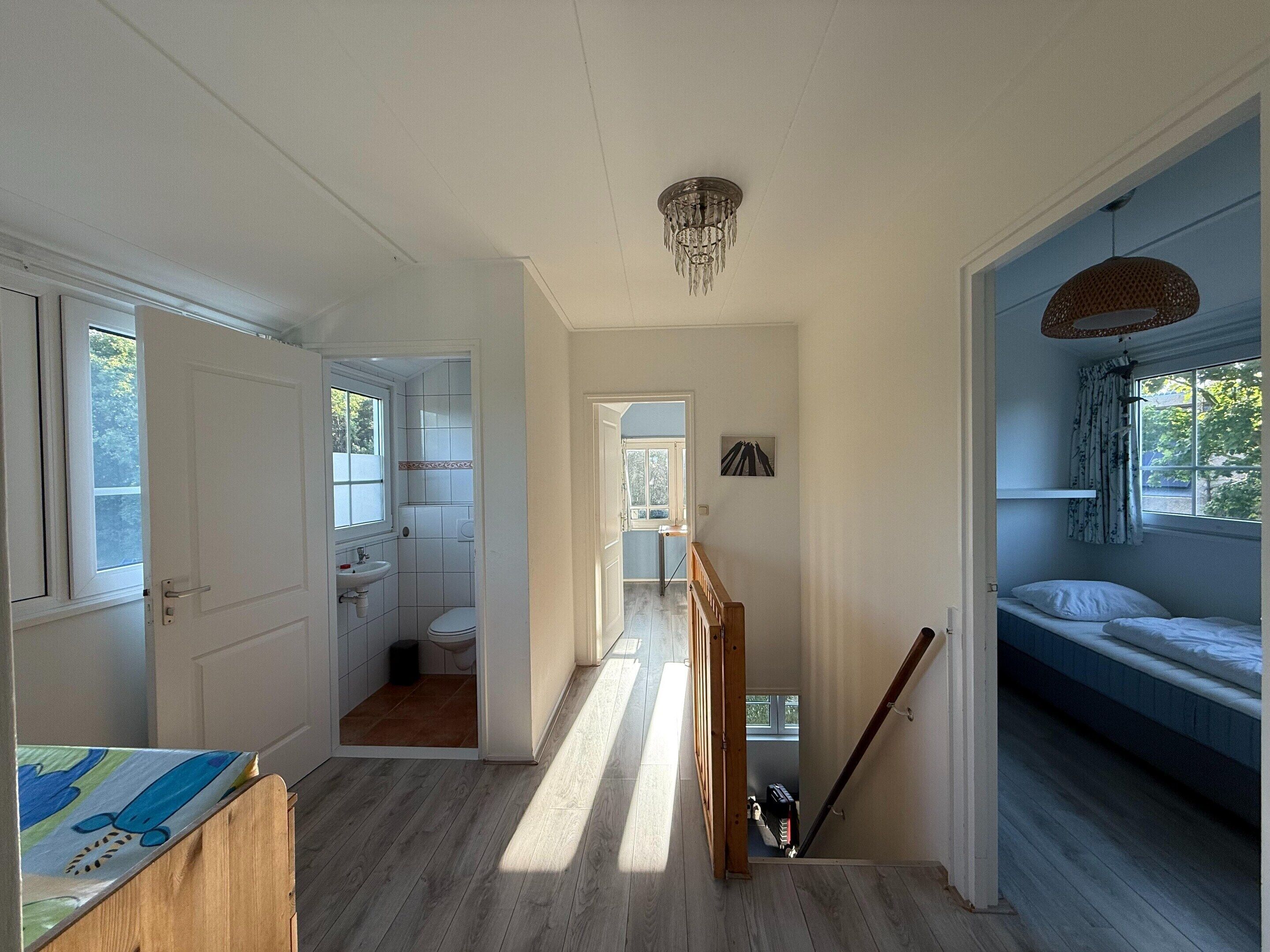 Een gang die leidt naar een slaapkamer met een stapelbed, een badkamer met toilet en wastafel, en een trap.