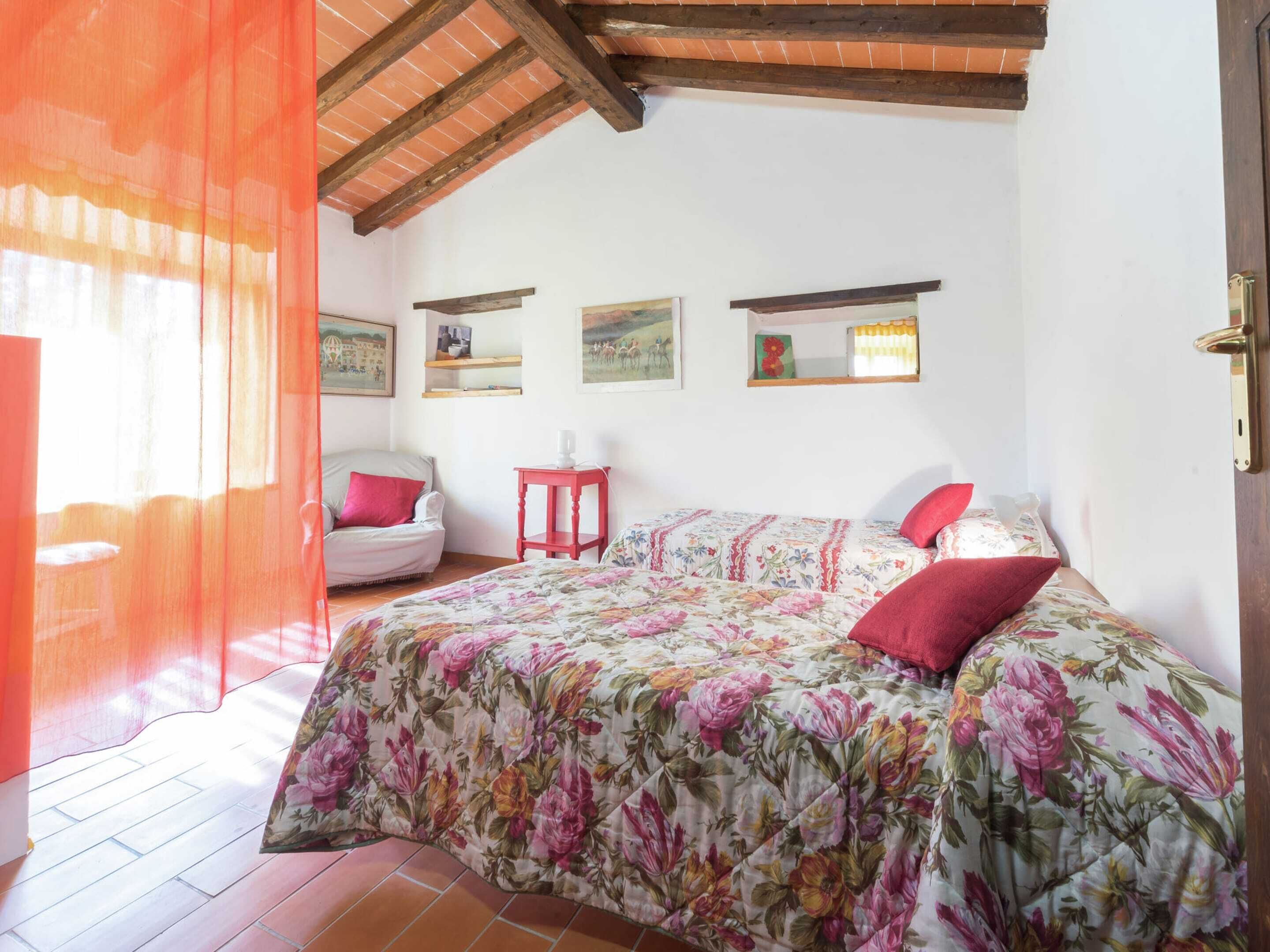 Quarto com edredom floral, uma cadeira, uma mesinha e obras de arte nas paredes.