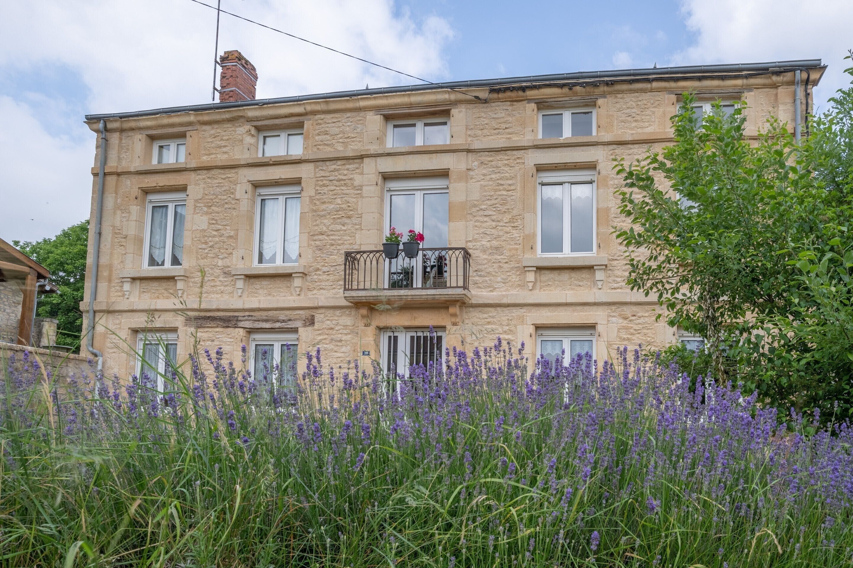 Un edificio de varios pisos con fachada de piedra, balcones con barandillas y un jardín con flores moradas.