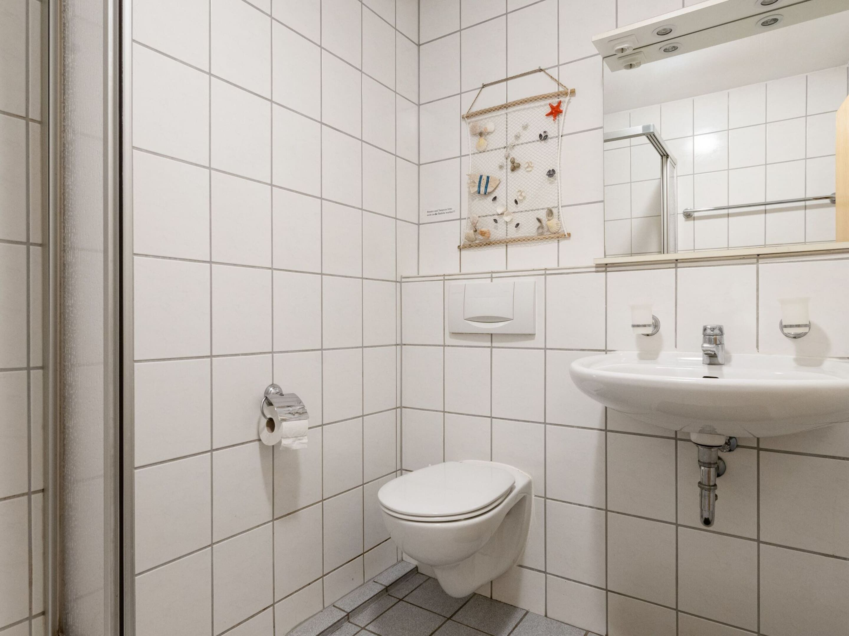 A small, clean bathroom with white tiles, a toilet, a sink, and a towel dispenser.