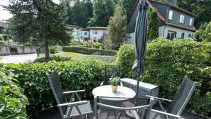 Eine Terrasse im Freien mit einem runden Tisch und vier Stühlen, umgeben von üppigem Grün und einer Hecke.