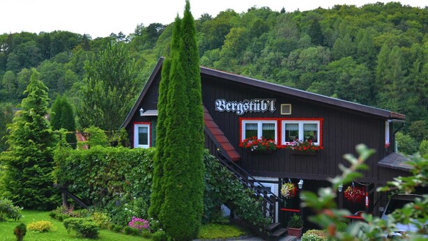 Ein traditionelles Holzhaus mit einem Schild, auf dem "Bergstübli" steht. Vor dem Eingang befindet sich eine Treppe, umgeben von üppigem Grün und einem gepflegten Garten.