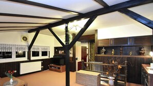 Ein gemütliches Wohnzimmer mit hohen Decken, freiliegenden Balken, einem Kamin, einem Fernseher und einer Küchenzeile mit Holzschränken und Dekorationselementen.