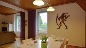 A dining area with a white table, chairs, and a green vase with flowers. There is a window with curtains, a sofa, and a wall decoration.
