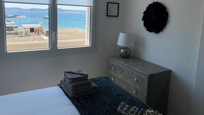 Une chambre avec un lit, une fenêtre donnant sur la mer, une lampe fixée au mur et un tableau encadré.