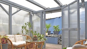 Une terrasse extérieure couverte, aménagée avec des meubles en osier, des plantes en pot et une table en verre.