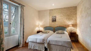 A room with two beds, a stone wall, a window with curtains, and a painting on the wall. - Elegant Manor Retreat Normandy (Ponts sur Seulles)