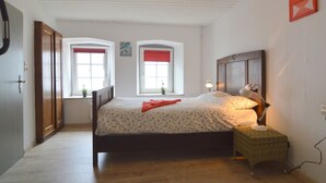 Ein Schlafzimmer mit einem hölzernen Bett, zwei Fenstern mit roten Vorhängen, einer Nachttischlampe und einem kleinen Tisch.