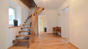 Eine Holztreppe führt zu einer zweiten Etage, ein Fenster mit einem Computer auf einem Regal und eine Tür am Ende des Flurs.