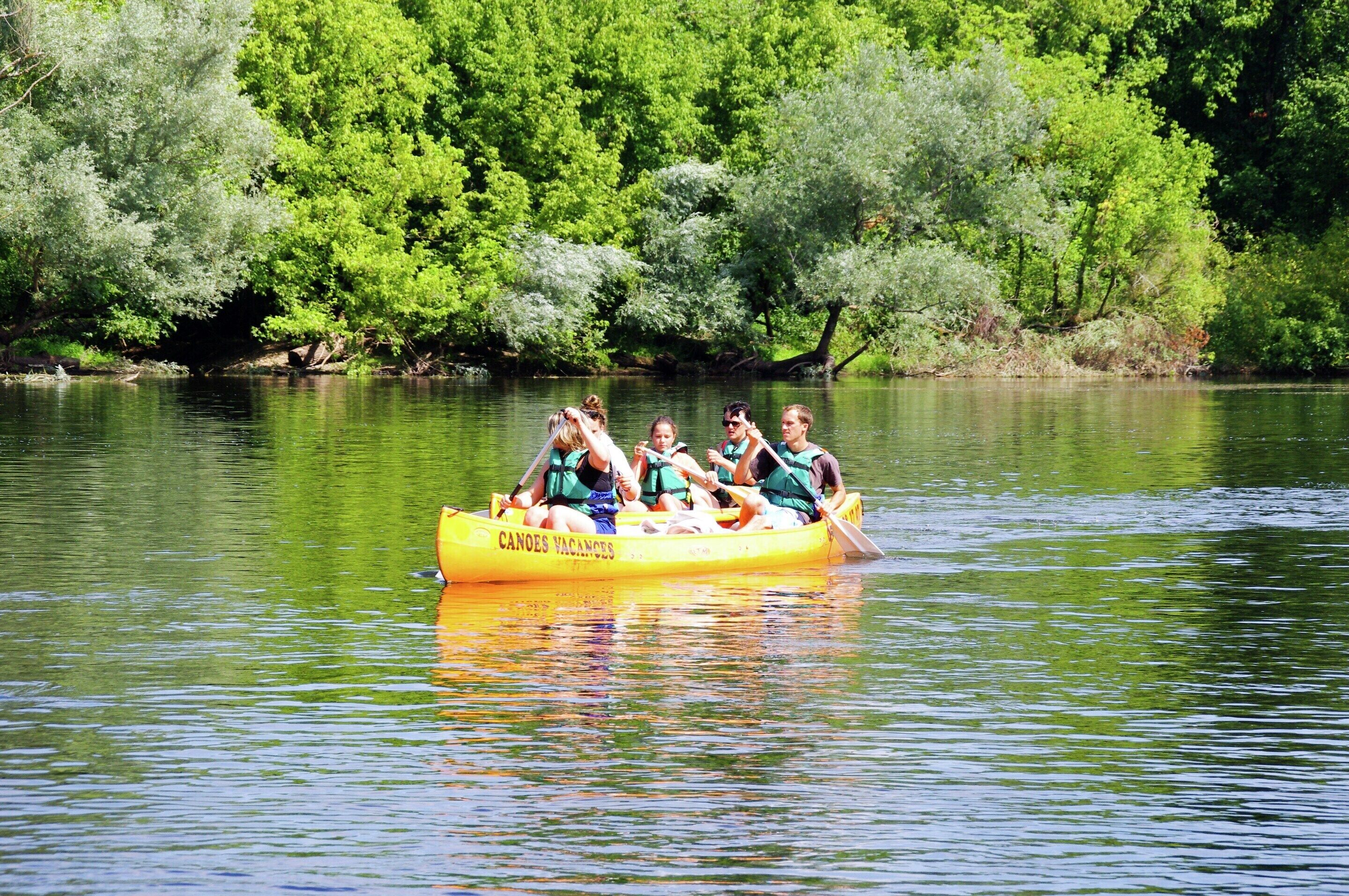 Una canoa amarilla con la leyenda "CANOE VACANCES", flotando en aguas tranquilas rodeada de frondosos árboles verdes.