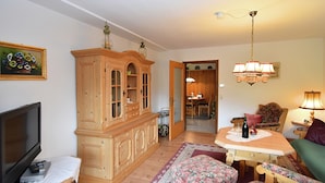 Una sala de estar con televisor, aparador de madera, otomana floral y una zona de comedor con mesa y sillas.