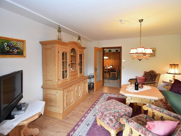 A living room with a television, wooden cabinet, floral ottoman, and a dining area with a table and chairs.