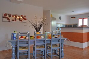 Un coin repas avec une table dressée pour quatre personnes, des chaises, un vase orné de branches et, au fond, une cuisine.