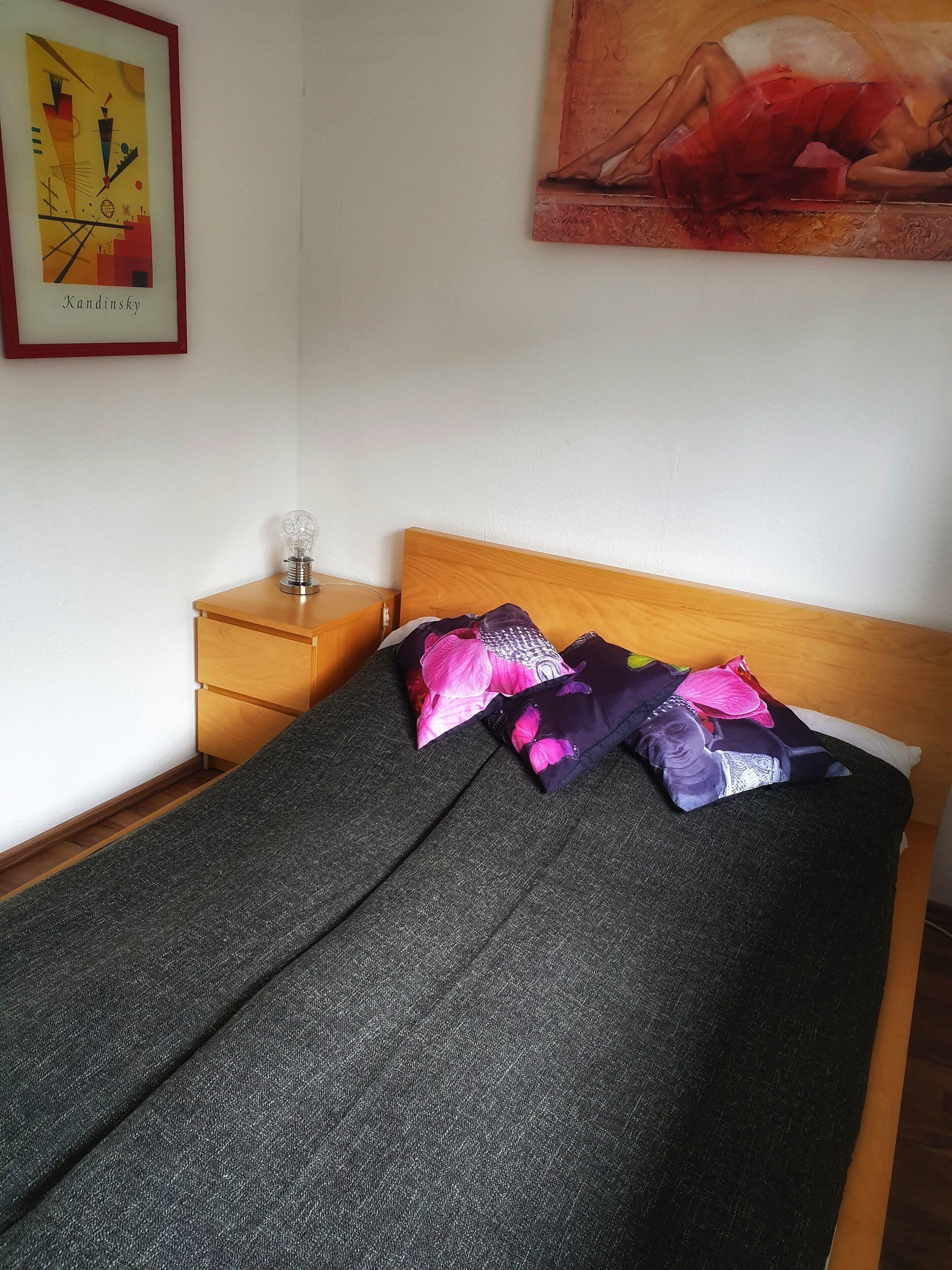Ein Schlafzimmer mit einem Bett, Kissen, einem Nachttisch, einer Lampe und einem Gemälde an der Wand.