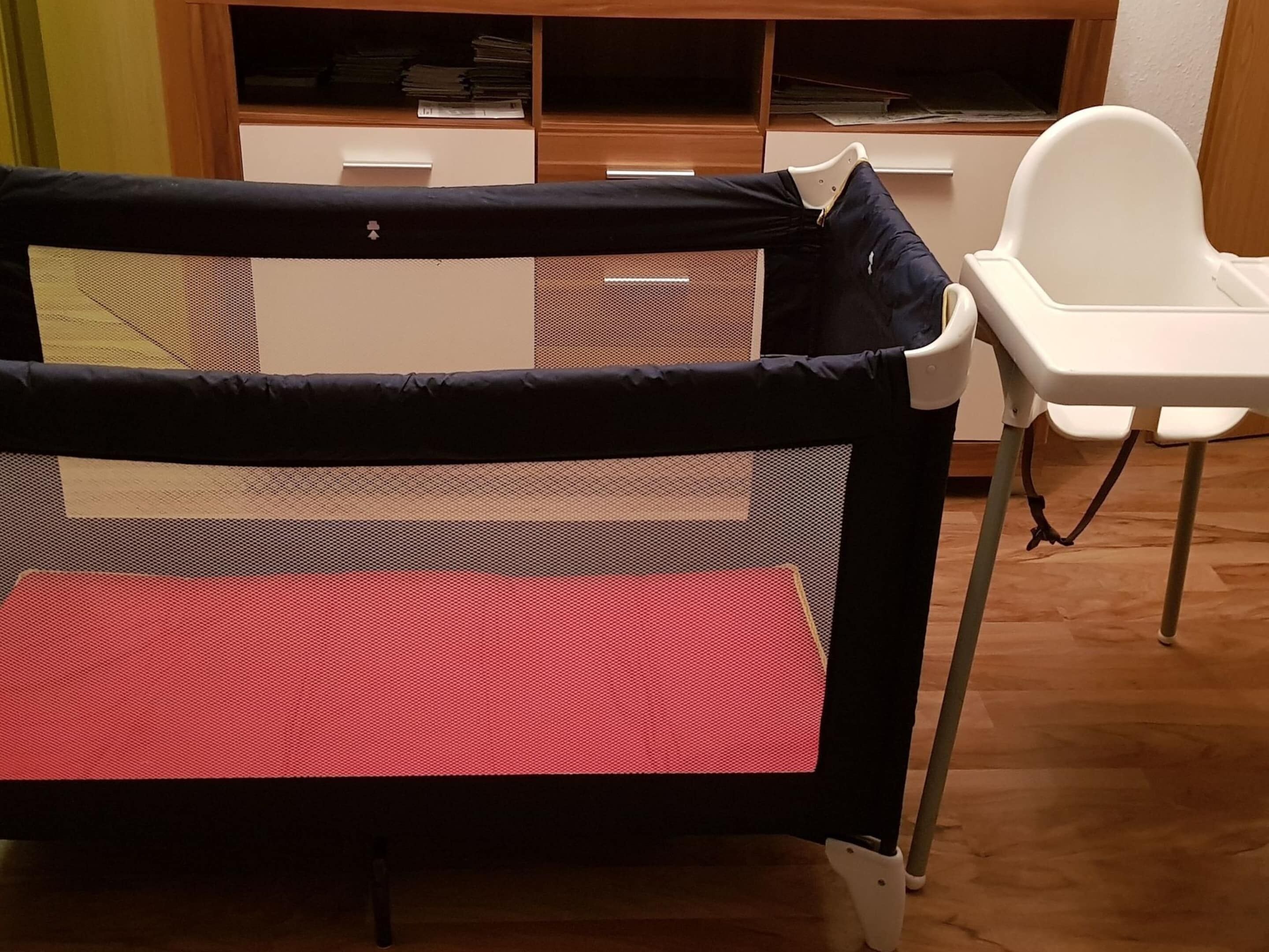 A room with a baby playpen, a white high chair, and a wooden cabinet with papers.