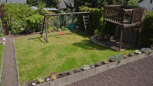A backyard with a swing set, a wooden playhouse, and a garden with flowers and plants. - Holiday Home in the Eifel With Balcony (Bremm)