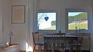 Ruokailutila, jossa on pöytä ja tuolit, sydämenmuotoisella koristeella varustettu ikkuna ja näkymä metsäiseen maisemaan.