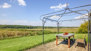 Une terrasse avec une table et un banc, entourée d’une pergola en métal, donnant sur un champ verdoyant et des arbres à l’horizon.