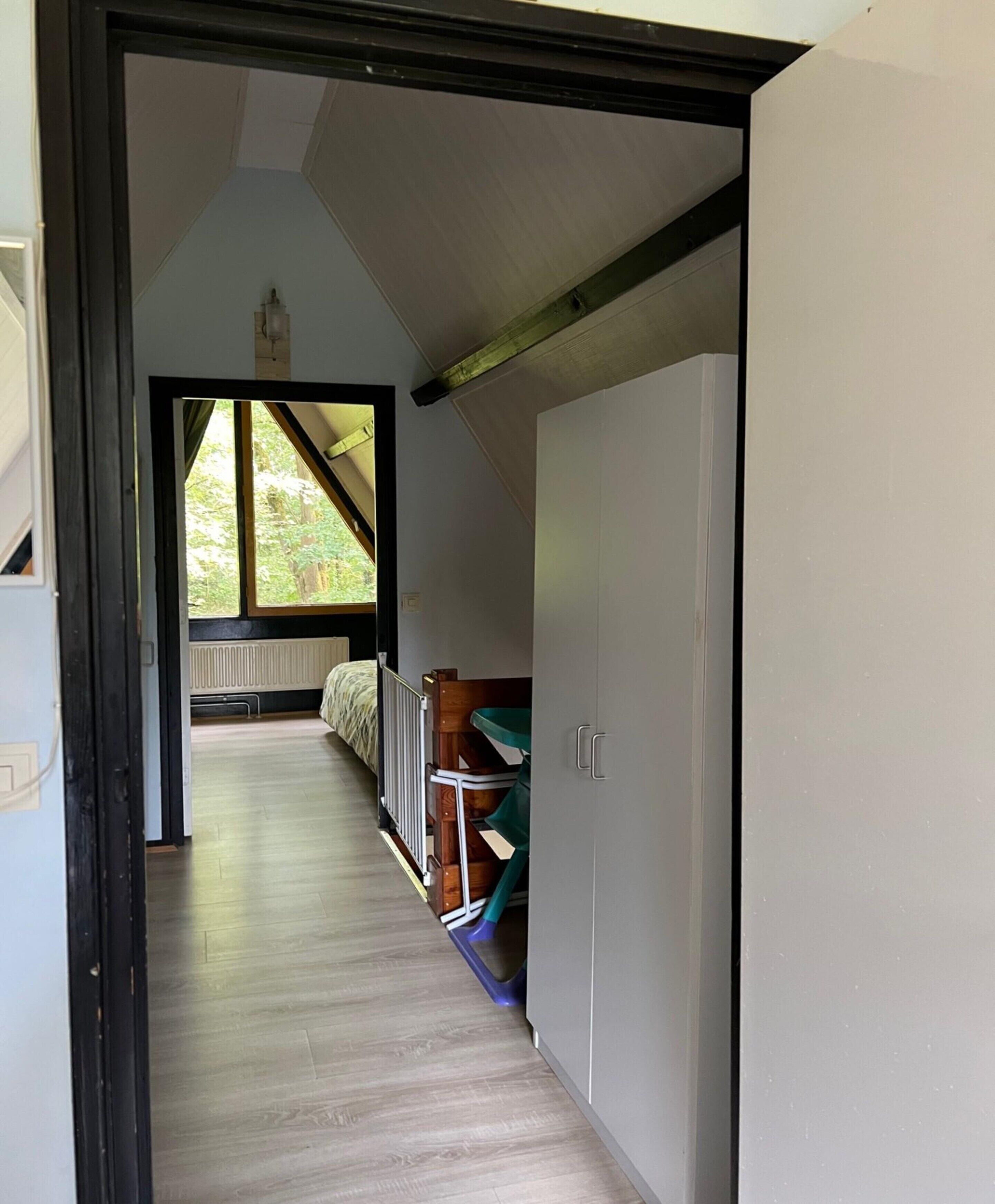 A narrow hallway with a wooden floor leading to a bedroom with a window, a radiator, and a bed.