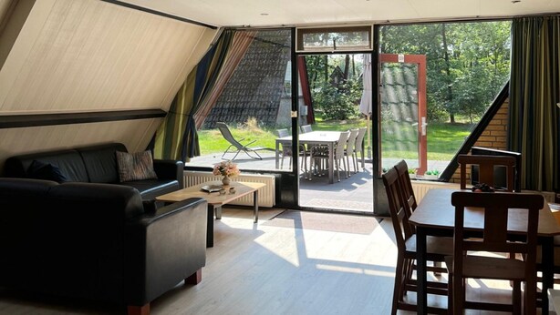 A modern living room with a black sofa, wooden coffee table, and a dining area with wooden chairs and a table. Large windows offer a view of the outdoors.