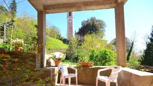A covered patio with two white plastic chairs, a potted plant, and a view of a clock tower in the background. - Holiday Home in Rio Near Fishing Villages (Sesta Godano)